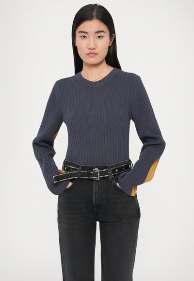 Camisola cinzenta canelada com mangas compridas e detalhes em mostarda, combinada com jeans pretos de cintura alta e um cinto preto com tachas.