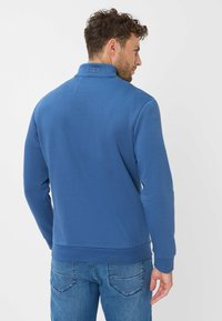 Pullover blu con colletto alto, polsini e orlo a costine. Presenta un logo in rilievo sul colletto e una texture liscia. Visibile un paio di jeans in denim.