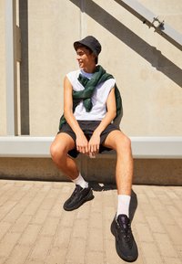 Ung man som bär en svart bucket hat, ärmlös vit skjorta, svarta shorts och sneakers, sitter på en kant mot en betongvägg.