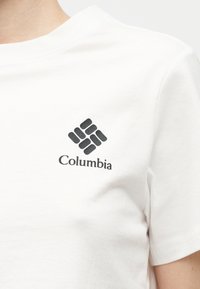 Λευκό πουκάμισο με κοντό μανίκι, με μαύρο λογότυπο Columbia και κείμενο στο αριστερό στήθος, φορεμένο από ένα άτομο μπροστά σε απλό φόντο.