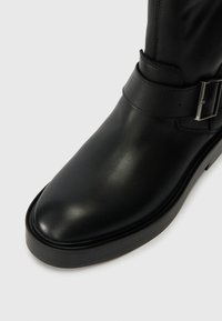 Bota de tornozelo em couro preto com uma textura suave, ponta arredondada e uma tira decorativa com detalhe de fivela prateada.