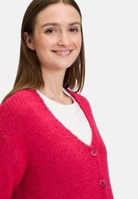 Fluffig rosa kofta med V-hals, prydd med två rosa knappar och ribbad textur, bärs över en vit skjorta. Närbild av plagget.