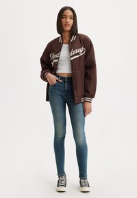 Chaqueta varsity marrón con letras blancas, cuello y puños de ribete, combinada con jeans ajustados azules y zapatillas de caña alta negras.
