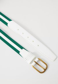 Vihreä ja valkoinen vyö, joka on valmistettu teksturoidusta kankaasta. Vyössä on valkoinen nahkaremmi ja kultainen metallinen solki, jossa on kohokuviointi.