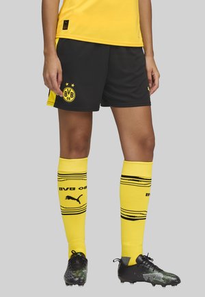 Pantalones cortos negros con acentos amarillos, que presentan un logo redondo. Medias de rodilla amarillas con rayas y logo negros. Zapatillas negras con acentos verdes.