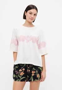 T-shirt blanche avec un imprimé floral rose sur la poitrine, manches courtes, associé à un short noir arborant des motifs floraux tropicaux colorés.