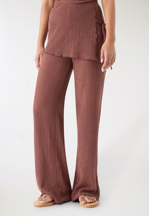 Donna che indossa pantaloni marroni a gamba larga con texture e un dettaglio di gonna avvolgente abbinata, abbinati a sandali infradito beige su uno sfondo bianco.