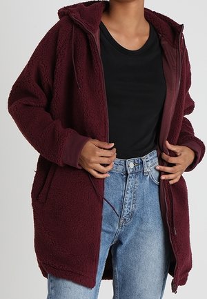 Persona vistiendo una sudadera con capucha de forro polar burdeos con cremallera sobre una camiseta negra y vaqueros de tiro alto azul claro, ajustando la sudadera con las manos.