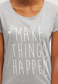 Camiseta gris con mangas dobladas que presenta el texto blanco "HAZ QUE LAS COSAS SUCEDAN" y dos gráficos de estrellas cerca del escote.