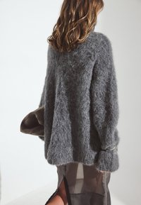 Übergroßer grauer Pullover mit flauschiger Textur, langen Ärmeln und silbernen Hardware-Details. Getragen über einem transparenten schwarzen Rock mit seitlichem Schlitz.