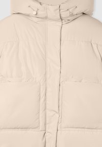 Kurtka puchowa w jasnym beżu z wysokim kołnierzem, sznurkiem do ściągania oraz dwiema dużymi kieszeniami z przodu; gładka, pikowana tekstura.