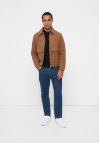 Giacca imbottita marrone con colletto, due tasche frontali, abbinata a un maglione nero e jeans blu. Sneakers bianche completano l'outfit.