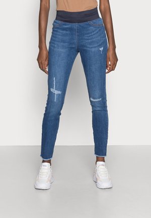 Person iført tætsiddende blå slidte jeans med en mørk elastisk taljebånd og hvide sneakers, stående mod en ensfarvet baggrund.