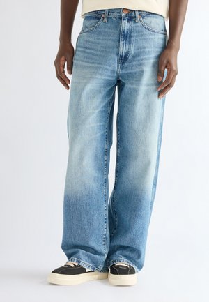 Person wearing light blue wide-leg jeans and black sneakers with white soles standing against a plain background.