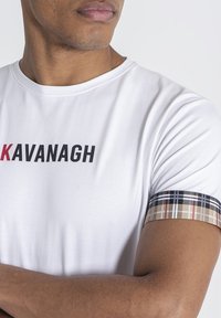 Camiseta blanca de manga corta con "KAVANAGH" impreso en negro y rojo. Presenta un patrón a cuadros en el puño de la manga en beige, negro y rojo.