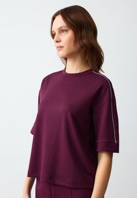 Bordeaux shirt met korte mouwen en een ronde halslijn, met een iets verlaagde schouderlijn. Voorzien van een zilveren accent langs de mouwnaad. Soepele stoftextuur.