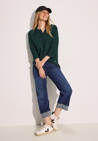 Haut V en textured vert foncé avec des manches trois quarts, associé à un jean bleu marine retroussé et des baskets blanches avec des accents noirs.