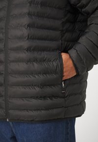 Sort isoleret jakke med horisontale quiltede detaljer, lynlåslommer og elastiske manchetter. Materialet er glat med en mat finish.