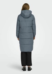 Manteau long matelassé gris avec capuche, présentant des coutures horizontales et des poignets élastiques, associé à un pantalon large noir et des chaussures noires.