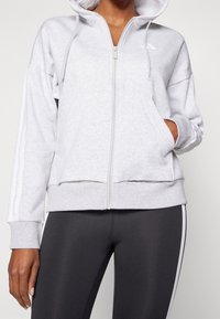 Grå zip-up hoodie med huva, som har två vita vertikala ränder på ärmarna. Mjuk textur, känguruficka och ribbade muddar.