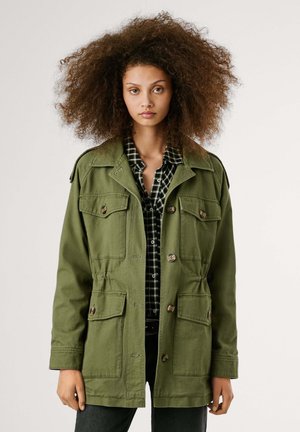Groene canvas jas met een kraag, meerdere voorzakken, knoopsluitingen en een verstelbare taille. Draagt over een zwart-wit geblokte shirt.