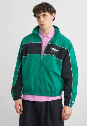 Jonge man met een groen-zwart Nike jack met rits over een roze poloshirt, staand tegen een effen grijze achtergrond.