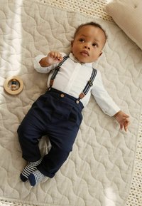 Dojenček v beli srajci z gumbi, temnomodri naramnicah, temnomodrih hlačah in s črtastimi nogavicami, ki leži na teksturirani bež odeji, poleg pa je lesena igrača.