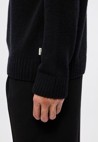 Vue de côté d'une personne portant un pull en laine noir avec des poignets et un ourlet côtes, montrant l'étiquette de la marque sur la couture latérale.