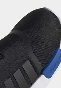 Svart idrottssko med en strukturerad tygöverdel, svarta gummidetaljer, blå logotyp på hälen och en vit sula.