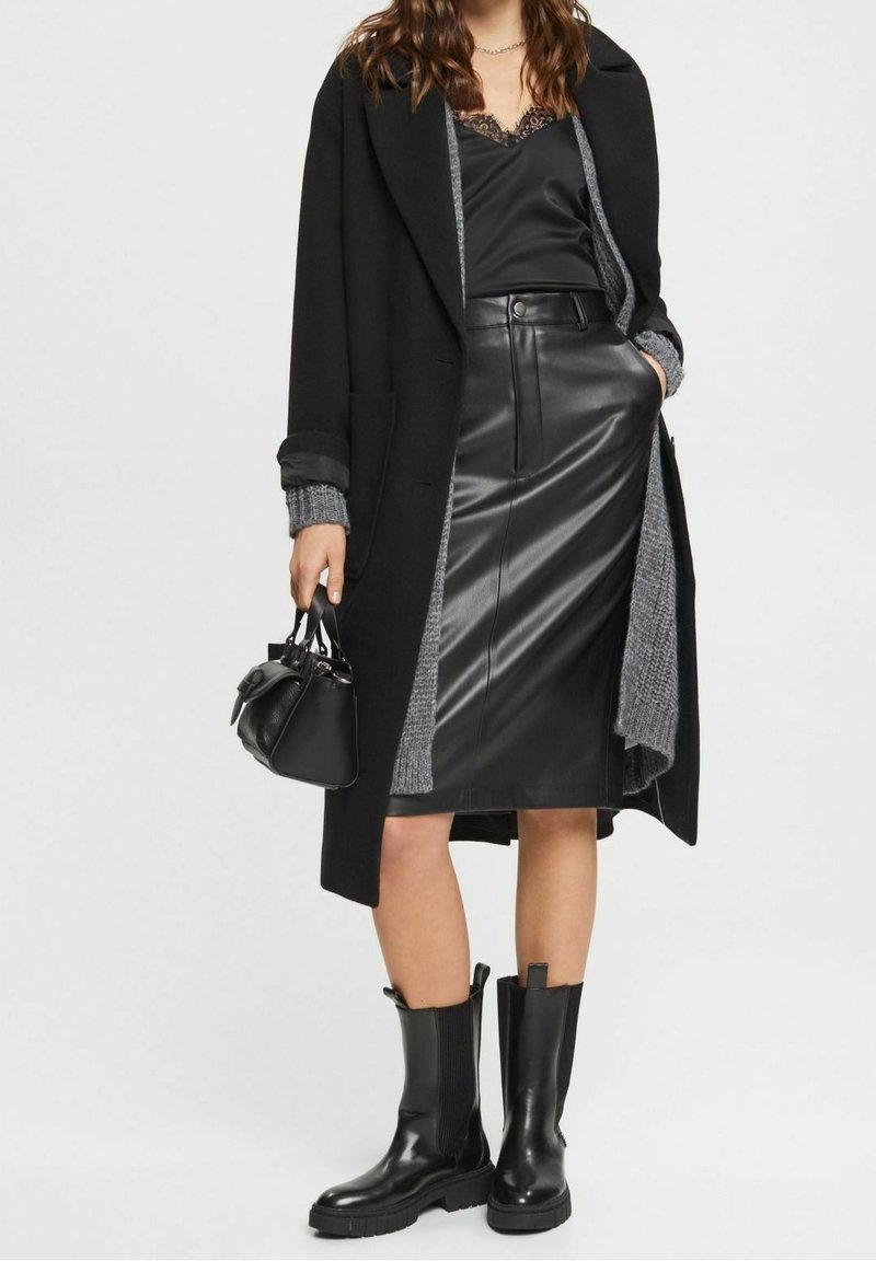 Femme portant une jupe en cuir noir, un caraco en satin noir, un long manteau noir avec des poignets en maille grise, tenant un sac à main noir et portant des bottines noires.