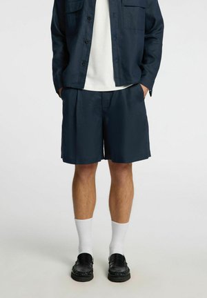 Pantaloni corti in lino blu navy con pieghe, abbinati a una camicia coordinata, una t-shirt bianca, calzini bianchi e scarpe slip-on nere.