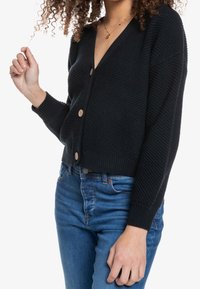 Cárdigan de punto negro con escote en V, adornado con botones de madera, dobladillos acanalados y un patrón texturizado, combinado con unos jeans de mezclilla azul.