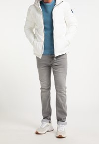 Weißer Pufferjacke mit Kapuze, Reißverschluss vorne und seitlichen Taschen, getragen über einem türkisfarbenen Strickpullover und grauen Jeans, kombiniert mit weißen Turnschuhen.