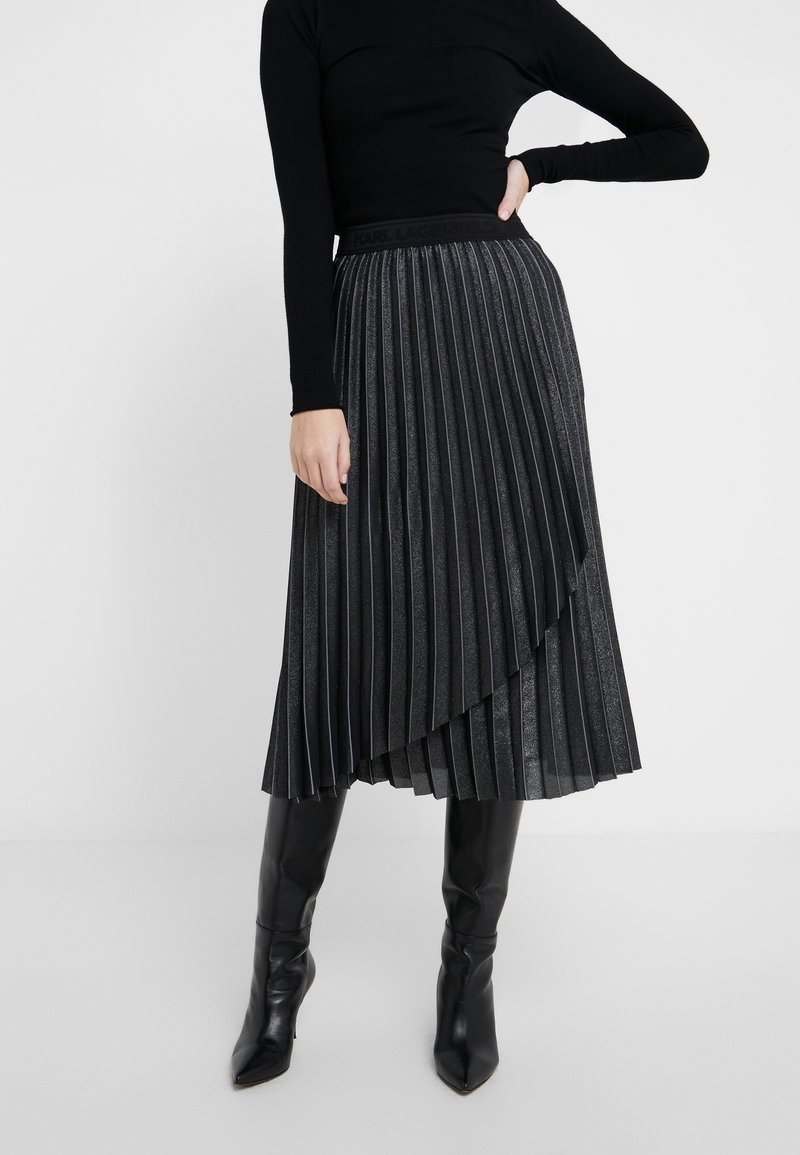 Falda midi plisada en tejido gris oscuro con un ligero brillo. Presenta un dobladillo asimétrico y una cinturilla negra con marca. Combinada con botas altas negras.