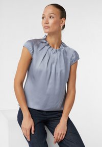 Himmelbluse mit kurzen Ärmeln, rüschigem Ausschnitt und gefaltetem Vorderteil. Der Stoff ist glatt und glänzend und ergänzt dunkle Hosen.