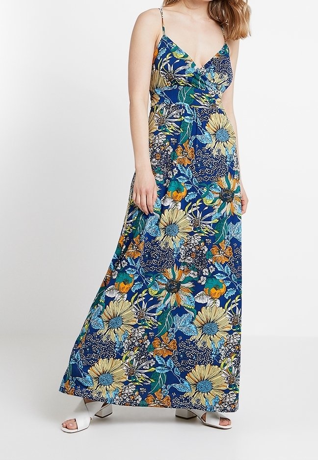 Femme portant une robe sans manches longue jusqu'aux chevilles avec des fleurs bleues, jaunes et oranges et des sandales blanches à bout ouvert.