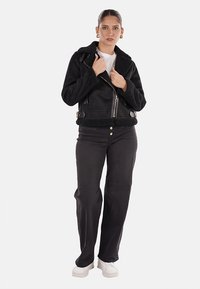Giacca bomber nera corta con colletto in pelliccia sintetica, chiusura zip e tasche con zip. Indossata con jeans neri e sneakers bianche.