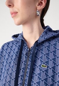 Huvtröja i marinblått med geometriskt mönster, dragkedjeförslutning och en liten grön Lacoste-logotyp på bröstet.