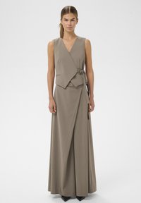 Kvinde iført en ærmeløs taupe omslagsvest og matchende bukser med vide ben, stående foran en ensfarvet, lys baggrund.