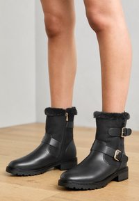 Bottines noires avec un bout en cuir et une tige en daim, ornées d'un revers en fausse fourrure, d'une fermeture éclair dorée et de deux sangles réglables.