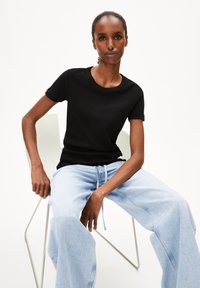 Sort kortærmet t-shirt lavet af ribbet stof, iført lyseblå løstsiddende jeans. Model siddende på en minimalistisk stol.