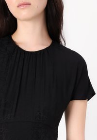 Black dress featuring soft fabric, a round neckline, short sleeves, and subtle embossed pattern. Pleats add texture and flow.