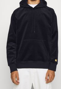 Marineblauer Hoodie aus weichem Material, mit einer großen Fronttasche, einer Kapuze mit Kordelzug und einem kleinen goldenen Logo am Ärmel.