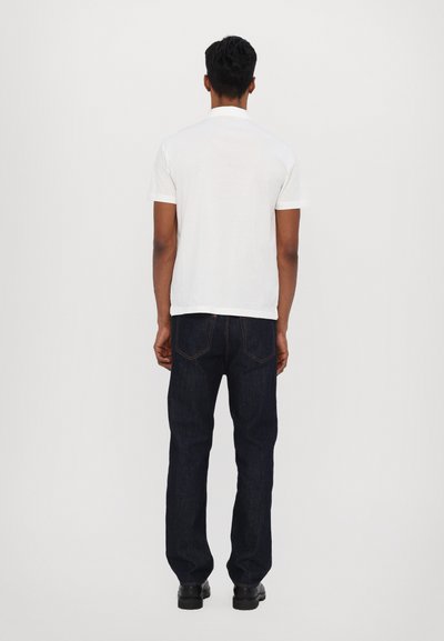 Balta trumpų rankovių polo marškinėliai derinami su tamsiai mėlynomis tiesiomis džinsomis. Marškiniai turi klasikinį apykaklę, o džinsai yra su užpakalinėmis kišenėmis.