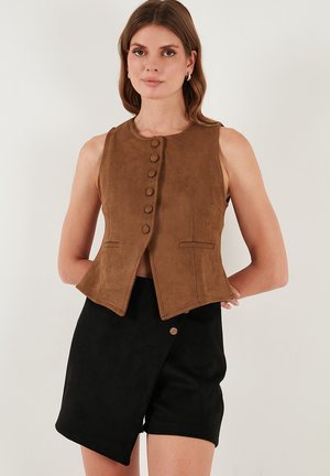 Barna suede mellény ujj nélküli dizájnnal, egy sor gombbal, oldalsó zsebekkel és elegáns, ferde alsó résszel. Fekete sorttal párosítva.