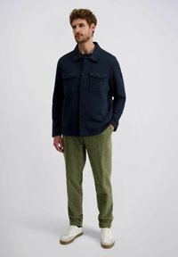 Donkerblauwe jas met twee borstzakken, knopen aan de voorkant en een kraag, gecombineerd met groene corduroy broek en witte sneakers.