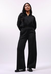 Zwarte zip-up top met een cropped ontwerp; wijde zwarte broek; gladde stof; gecombineerd met zwarte schoenen met witte strepen.