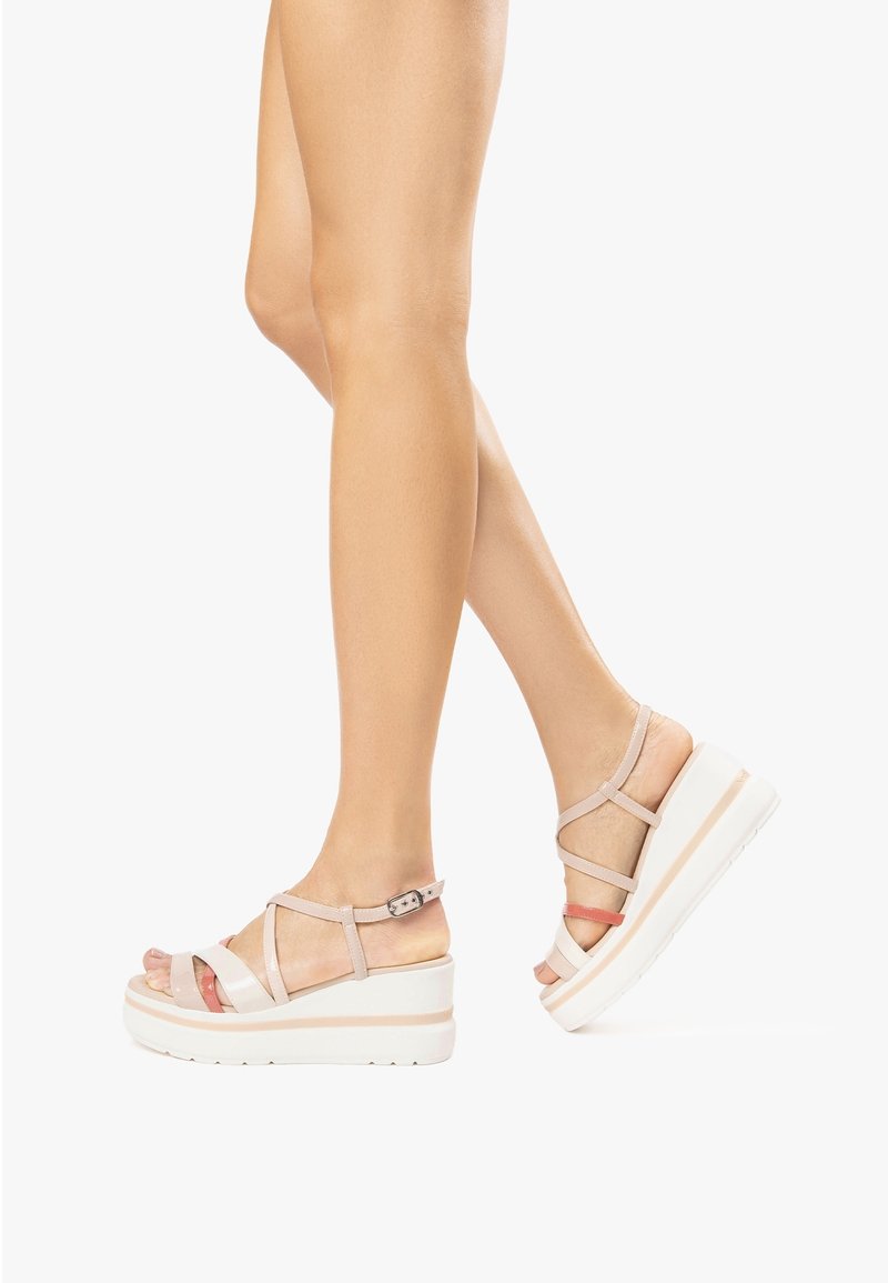 Jambes portant des sandales compensées beiges à plate-forme avec brides à la cheville, en train de faire un pas sur un fond blanc.