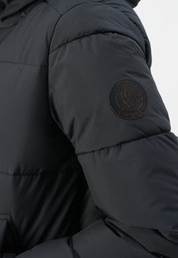Zwarte gewatteerde jacksleeve met een cirkelvormige patch die een eend toont en de tekst "ARCTIC SAVE THE DUCK" op de bovenarm.