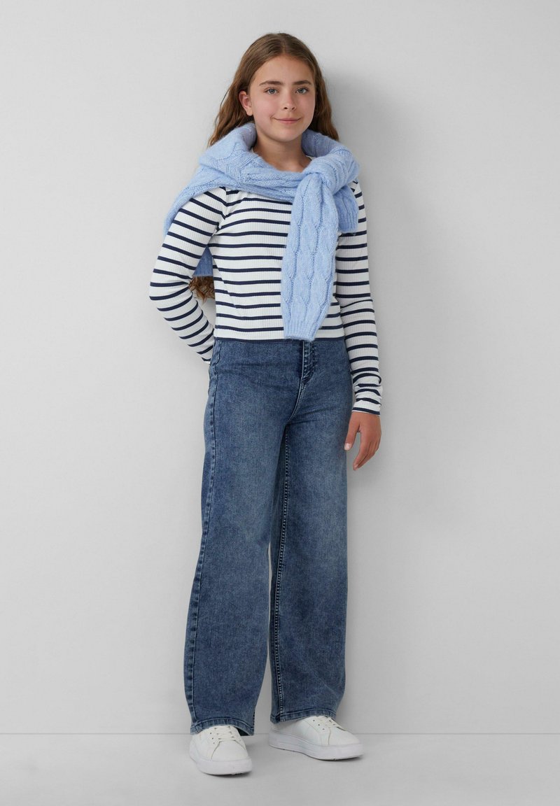 Pruhovaná dlhá košeľa s dlhým rukávom v námorníckej modrej a bielej farbe, v kombinácii s vysokými modrými džínami a svetlomodrým pleteným šálom. Biele tenisky.